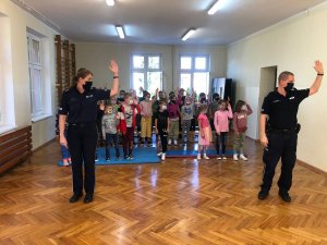 Policjant i policjantka stoją tyłem do grupy dzieci  w dużych odstępach.  Mają uniesione w górze lewe ręce. Dzieci stoją i naśladują zachowania policjantów (trzymając podniesioną w górze lewą rękę) ucząc się prawidłowego rozglądania przy przechodzeniu prze jezdnię czy przejściu dla pieszych. Zajęcia odbywają się w sali gimnastycznej.