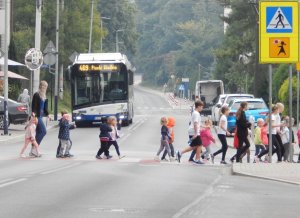 Przejście dla pieszych oznakowane znakiem drogowym pionowym „przejście dla pieszych” wraz z tabliczką „agatka” wskazującą że przejście dla pieszych jest szczególnie uczęszczane przez dzieci. Na przejściu znajduje się grupa przedszkolna wraz z opiekunami. Dzieci mają podniesione dłonie, (gest ręką by zwrócić uwagę kierujących na to że ktoś przechodzi przez jezdnię). Do przejścia podjeżdża autobus miejski z wyświetlonym napisem w górnej części pojazdu – „469 PIASKI WIELKIE”. W tle perspektywa ulicy w obszarze zabudowanym. Umiarkowany ruch, dobre warunki drogowe.