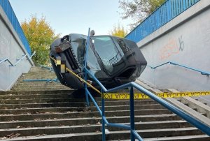 Samochód koloru czarnego oparty na barierkach w przejściu podziemnym dla pieszych. Auto oparte na boku. Widać uszkodzone barierki.