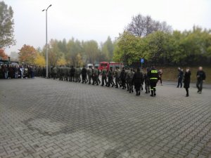 Pokaz musztry paradnej uczniów Zespołu Szkół w Olkuszu ZDZ przed budynkiem Miejskiego Ośrodka Sportu i Rekreacji w Olkuszu. Pogoda pochmurna w tle widać wóz strażacki oraz zgromadzonych widzów.