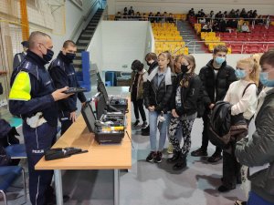 Stanowisko wystawowe policjantów Komendy Powiatowej Policji w Olkuszu. Policjant w Odblaskowej kamizelce rozmawia z osobami gromadzącymi się przed stoiskiem i pokazuje sprzęt będący na wyposażeniu policjanta ruchu drogowego. Policjant trzyma w ręce urządzenie mobilne. W tle trybuny i uczniowie na trybunach.