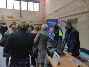 Stanowisko wystawowe policjantów Komendy Powiatowej Policji w Olkuszu. Policjant w odblaskowej kamizelce rozmawia z osobami gromadzącymi się przed stoiskiem i pokazuje sprzęt będący na wyposażeniu policjanta ruchu drogowego. Obok stoi dzielnicowy.  Policjant ruchu drogowego  trzyma w ręce alcosensor, urządzenie do badania stanu trzeźwości kierujących.  W tle logo i adres Komendy Powiatowej Policji w Olkuszu.