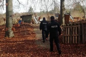 Zdjęcie przedstawia dwóch umundurowanych policjantów, którzy ścieżką idą w stronę baraku. Wokół liście i drzewa. pogoda jesienna.