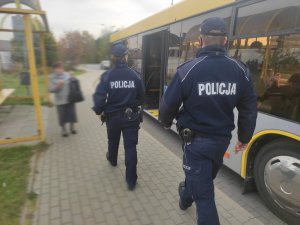 Policjantka i policjant idą w stronę autobusu. Po prawej znajduje się zatoka autobusowa i  żółty autobus. Po lewej stronie, przystanek autobusowy,  obok stoi kobieta. W tle widać drogę. Pogoda jesienna.