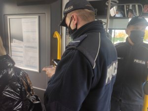 Dwóch umundurowanych policjantów podczas legitymowania osoby. Funkcjonariusze znajdują się w autobusie. Jeden z nich trzyma w ręce dowód osobisty.