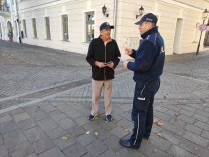 Policjant wręcza ulotkę mężczyźnie i rozmawia z nim o zagrożeniach. W tle widać olkuski Rynek i zabudowania.