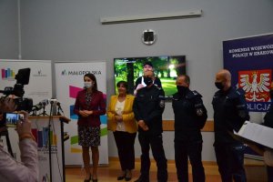 Przedstawiciele urzędu marszałkowskiego oraz policjanci podczas konferencji prasowej &amp;quot;Wnuczek, wnuczka edukuje - babcia, dziadek się stosuje&amp;quot;. osoby udzielają wywiadu do prasy. W tle plakaty Wojewódzkiego Urzędu Marszałkowskiego oraz Komendy Wojewódzkiej Policji w Krakowie.