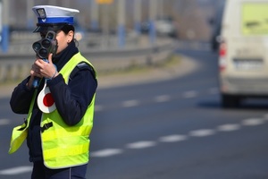 Policjantka ruchu drogowego (widoczna żółta kamizelka, biała czapka) mierzy prędkość ręcznym miernikiem. W tle  - ruch uliczny.