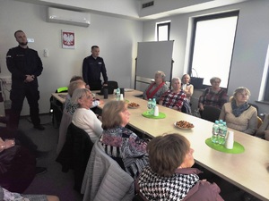 Komendant Komisariatu Policji oraz dzielnicowy podczas prelekcji z seniorami, znajdują się w auli na komisariacie, gdzie prowadzone sa spotkania i narady. Na około stoły i siedzące za nimi kobiety, uczestnicy  spotaknia.