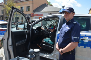 Dziecko siedzi za kierownica radiowozu. Obok stoi policjant. W tle widać zabudowania i olkuski Rynek. Pogoda letnia. Słoneczne niebo.