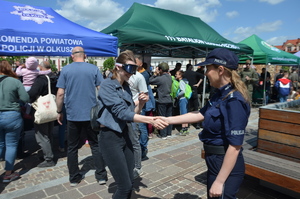 Kobieta w narkogoglach podaje dłoń policjantce. W tle namioty widać stanowisko  żołnierzy i policjantów oraz gromadzących  się wokół nich ludzi.