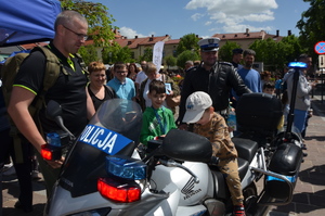 Chłopiec siedzi za kierownicą policyjnego motocyklu. W tle policjant ruchu drogowego oraz gromadzący się ludzie. Pogoda letnia. Słoneczne niebo.