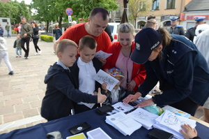 Policjantka wykonuje dzieciom odciski palców na dyplomie. W tle olkuski rynek. Pogoda letnia. Słoneczne niebo.