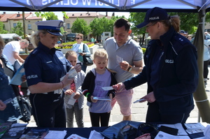 Policjantka ruchu drogowego (biała czapka, biały pas) wraz z policjantka prewencji rozdają ulotki osobom zgromadzonym przy stanowisku policyjnym.