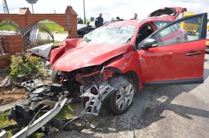 Czerwone Renault po wypadku. Przód samochodu doszczętnie zniszczony. Na poboczu drogi widać porozrzucane elementy karoserii.W tle uszkodzone ogrodzenie przy posesji oraz służby.