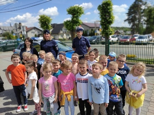 Policjantka prewencji i policjant ruchu drogowego (biała czapka) z przedszkolakami pozują do zdjęcia. W tle widać drogę oraz zabudowania. Pogoda letnia słoneczna.