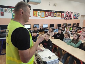 Technik kryminalistyki w odblaskowej kamizelce prowadzi zajęcia w klasie. Dzieci siedzą w szkolnych ławkach.