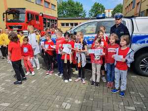 Policjant ruchu drogowego z dziećmi przy radiowozie pozują do zdjęcia. Dzieci trzymają w rękach dyplomy z własnymi odciskami linii papilarnych.