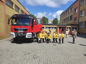 Wóz strażacki, przy którym stoi grupa dzieci. W tle budynek szkoły. Pogoda słoneczna letnia.