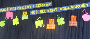 Na niebieskim tle znajduje się napis &quot;bądź bezpieczny i zdrowy, noś element odblaskowy&quot;. Poniżej odblaski o różnych kształtach i kolorach.