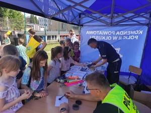 Policjanci robią dzieciom odciski palców w namiocie policyjnym.  Wokół stanowiska profilaktycznego gromadzą się dzieci. W tle widać boisko szkolne.