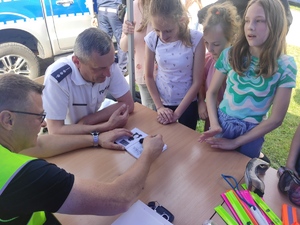 Technik kryminalistyki przy stanowisku profilaktycznym przy szkole pokazuje dzieciom, jak ujawnić ślady palców na papierze. Obok siedzi policjant ruchu drogowego w białej koszulce.