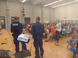 Dwaj umundurowani policjanci prowadza zajęcia. Obok policjantów widać projektor. Zgromadzone licznie dzieci siedzą w sali konferencyjnej.