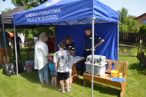 Piknik w Małobądzu. Namiot profilaktyczny policji, na stoliku znajdują się ulotki, gadżety i kolorowe broszury. Kobieta z dziećmi stoi przy stanowisku, naprzeciwko policjantka i policjant rozmawiają z dziećmi. Pogoda letnia, słoneczna.