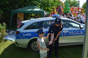 Piknik w Małobądzu. Chłopiec w policyjnej czapce przybija piątkę policjantce. Z tyłu oznakowany radiowóz. W tle grupa ludzi. Pogoda letnia, słoneczna.