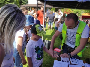 Piknik w Małobądzu. Technik kryminalistyki przy stanowisku policyjnym wykonuje odciski linii papilarnych. W tle widać ludzi. Pogoda letnia, słoneczna.