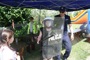 Chłopiec ubrany w kamizelkę przeciwuderzeniową ,ma na głowie hełm, w ręku trzyma tarcze i pałkę służbową obok stoi Komendant Komisariatu Policji w Kluczach. W tle kobieta w odblaskowej kamizelce z napisem Policja.