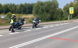 Policjant ruchu drogowego dokonuje  pomiaru prędkości. Obok policyjny motocykl. W tle widać drogę. Pogoda letnia, niebo bezchmurne.