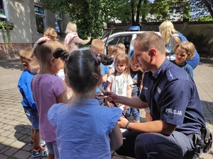 Dzielnicowy kuca przy przedszkolakach i pokazuje im sprzęt i wyposażenia policjanta. Wokół niego gromadzi się kilkuosobowa grupa przedszkolaków.