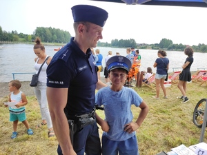 Chłopiec w policyjnej czapce pozuje do zdjęcia z policjantem przy stanowisku policyjnym. W tle Zalew Wolbromski i gromadzące się przy zalewie osoby w kamizelkach ratunkowych.