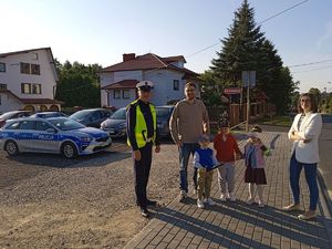 Policjant Ruchu Drogowego stoi na chodniku z rodzicami i ich trójką dzieci dwoma chłopcami i dziewczynką trzymaja oni odblaski w tle parking samochodowy z zaparkowanym radiowozem zabudowania drzewa ulica słoneczna pogoda