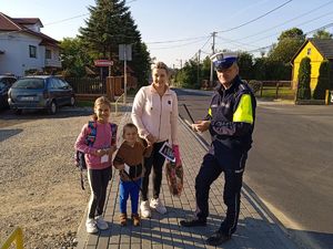 Policjant Ruchu Drogowego stoi na chodniku z matka z dwójką dzieci chłopczykiem i dziewczynką wręcza im elementy odblaskowe w tle parking samochodowy, zabudowania i ulica słoneczna pogoda