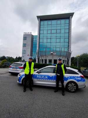 Dwóch policjantów Ruchu Drogowego stoi przy radiowozie. W tle budynek  Komendy Powiatowej Policji w Olkuszu. Pochmurna pogoda.