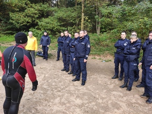 Grupa umundurowanych  policjantów podczas szkolenia wodnego. Na zdjęciu po lewej stronie stoi   ratownik wodny w kombinezonie nurka. W tle widać las i drzewa .