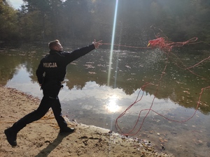Policjant w ruchu nad brzegiem wody rzuca rzutką do wody. W tle widać drzewa i las. Pogoda jesienna słoneczna.