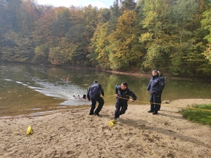 Dwaj policjanci wyciągają z wody pozoranta ciągnąc żółtą linę rzutki. Po prawej stoją dwaj  komendanci. W tle widać las. Pogoda jesienna słoneczna.
