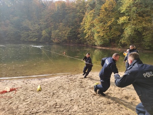 Trzech umundurowanych policjantów przy pomocy liny wyciąga na brzeg osobę,  znajdująca się w wodzie. Postawa policjantów w ruchu, akcja dynamiczna.   Obok po prawej stronie stoją dwaj komendanci. W tle widać drzewa. Pogoda jesienna, słoneczna .