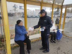 Policjantka prewencji podaje starszej Pani ulotkę. Kobieta siedzi na przystanku autobusowym. W tle widać sklep i zaparkowane samochody. Pogoda pochmurna, jesienna.