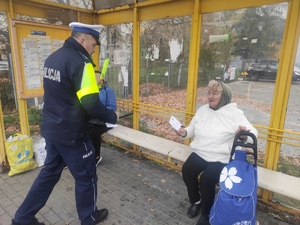 Policjant ruchu drogowego podchodzi do kobiety, która siedzi na przystanku autobusowym. Policjant przekazuje seniorce  ulotkę. W tle budynki, parkin. Za przystankiem autobusowym widać na trawie i parkingu  kolorowe liście. Pogoda jesienna.