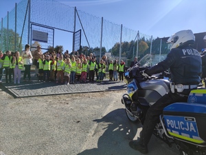 Po prawej na zdjęciu policjant ruchu drogowego na motocyklu, w białym kasku. Przed nim przodem stoją dzieci w odblaskowych kamizelkach oraz  nauczyciele. Za dziećmi widać boisko szkolne. Pogoda słoneczne, jesienna. Niebo błękitne.