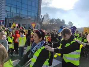 Dzieci w odblaskowych kamizelkach trzymają się za ramiona (jeden z drugim). Część dzieci tańczy. Pozostała grupa stoi przed budynkiem Komendy Powiatowej Policji w Olkuszu. Pogoda jesienna, słoneczna.  Bezchmurne niebo. W tle widać zabudowania oraz drzewa.