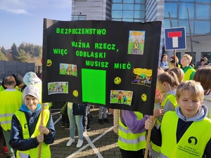 Uczniowie w odblaskowych kamizelkach przed budynkiem Komendy Powiatowej Policji w Olkuszu. Dwóch uczniów trzyma w ręce transparent z odblaskami i hasłem „Bezpieczeństwo ważna rzecz, więc odblaski musisz mieć”. Pogoda jesienna, bezchmurne niebo.