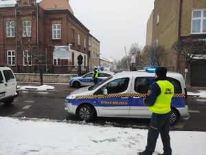 Funkcjonariusz straży miejskiej w odblaskowej kamizelce stoi przy samochodzie służbowym, który ma włączone sygnały świetlne. W tle widać policjantów ruchu drogowego przy radiowozie. Budynek szkoły, bramę wjazdowa i zaparkowane samochody. Zdjęcie zrobione pora zimową.