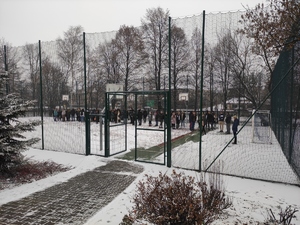 Grupa młodzieży ewakuowanej ze szkoły, stoi w miejscu wyznaczonym, poza budynkiem szkolny. Zimowa pora. Na boisku widać śnieg.