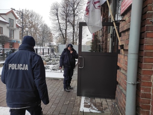 Dwaj policjanci wchodzą do budynku szkoły. Jeden trzyma w ręce radiostację i porozumiewa się z dowodzącym akcją.