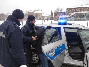 13.	Zatrzymany wprowadzany jest do radiowozu przez dwóch policjantów. W tle olkuski Rynek i zabudowania. Pora zimowa.